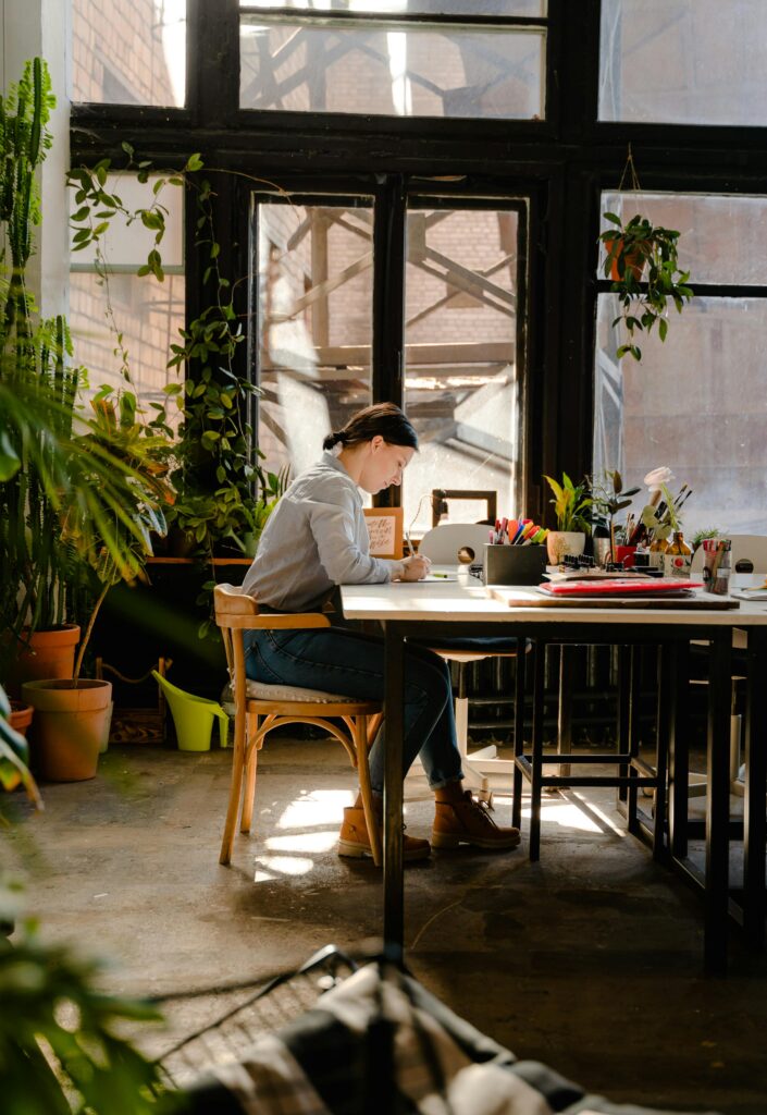 pexels-photo-4348078-4348078 A female artist working diligently in a sunlit studio surrounded by plants and art supplies.